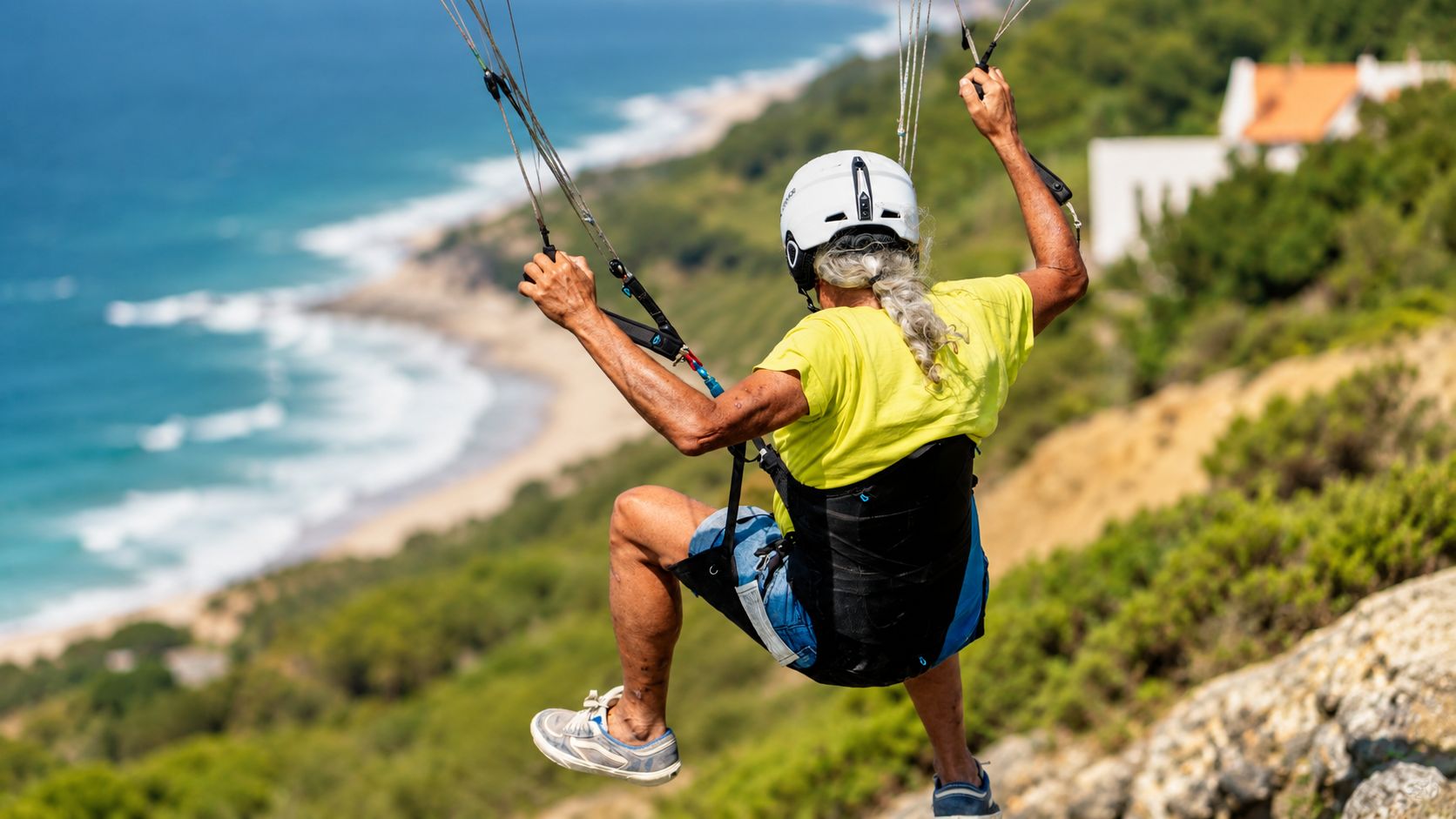 Piloto a descolar de uma encosta sobre a costa, com as duas mãos nos comandos e o harness à vista.