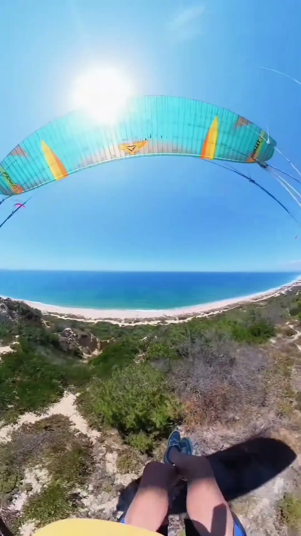 A vista do piloto sob um parakite Flow turquesa, em voo baixo sobre a vegetação das dunas da Fonte da Telha, com a praia e o Atlântico ao fundo.