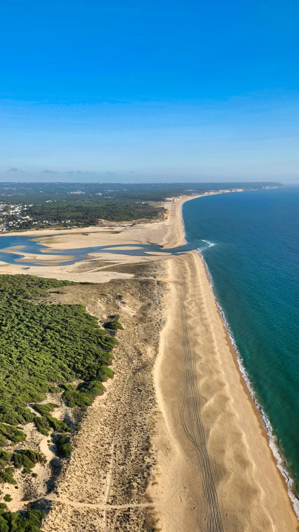 Vista aérea da Lagoa de Albufeira: a lagoa com bancos de areia à esquerda, o pinhal, a praia extensa e o mar aberto à direita.