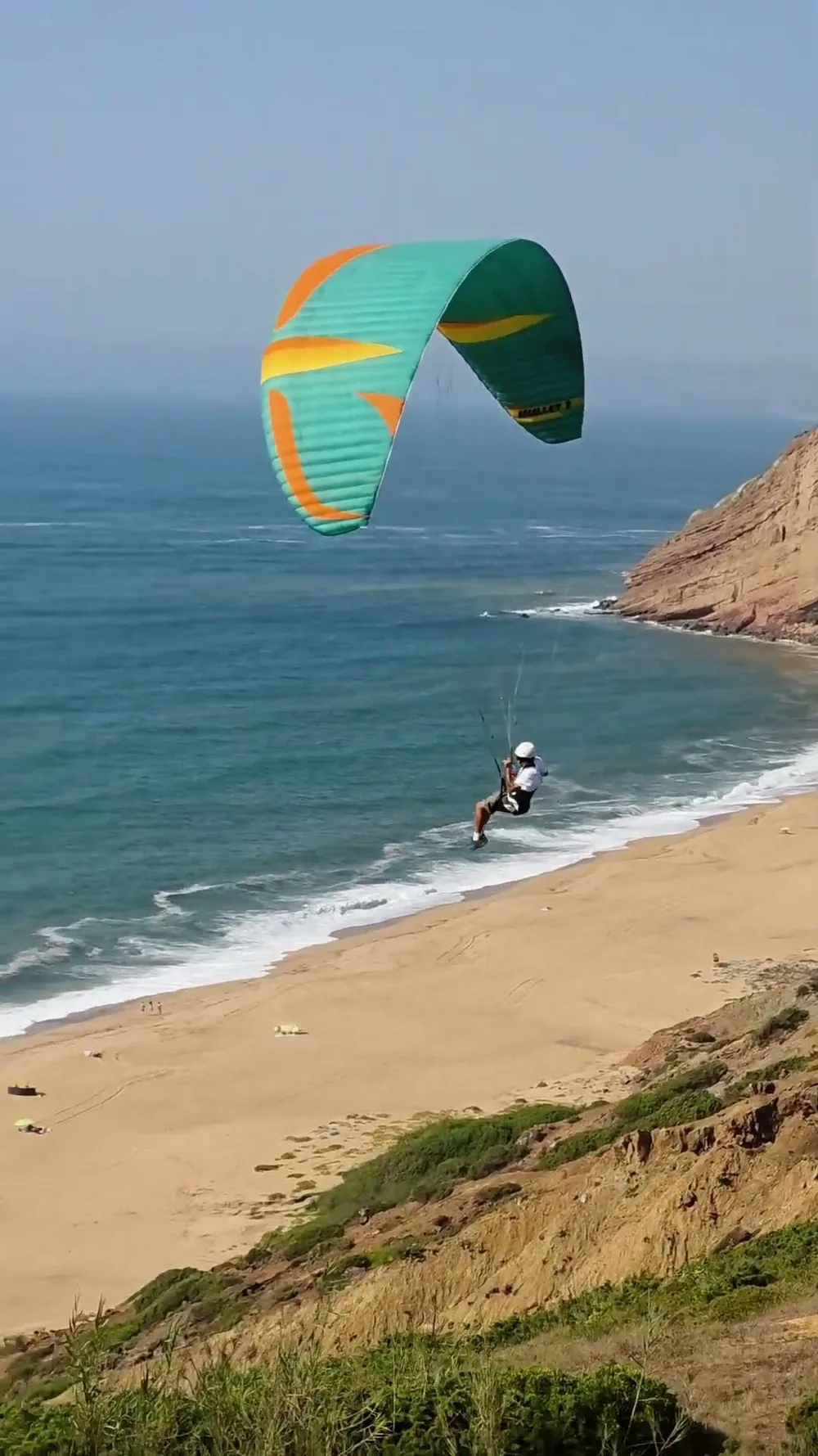 Um parakite turquesa e laranja a voar baixo sobre o areal da Praia da Gralha, entre o Atlântico à esquerda e as arribas ocres à direita.