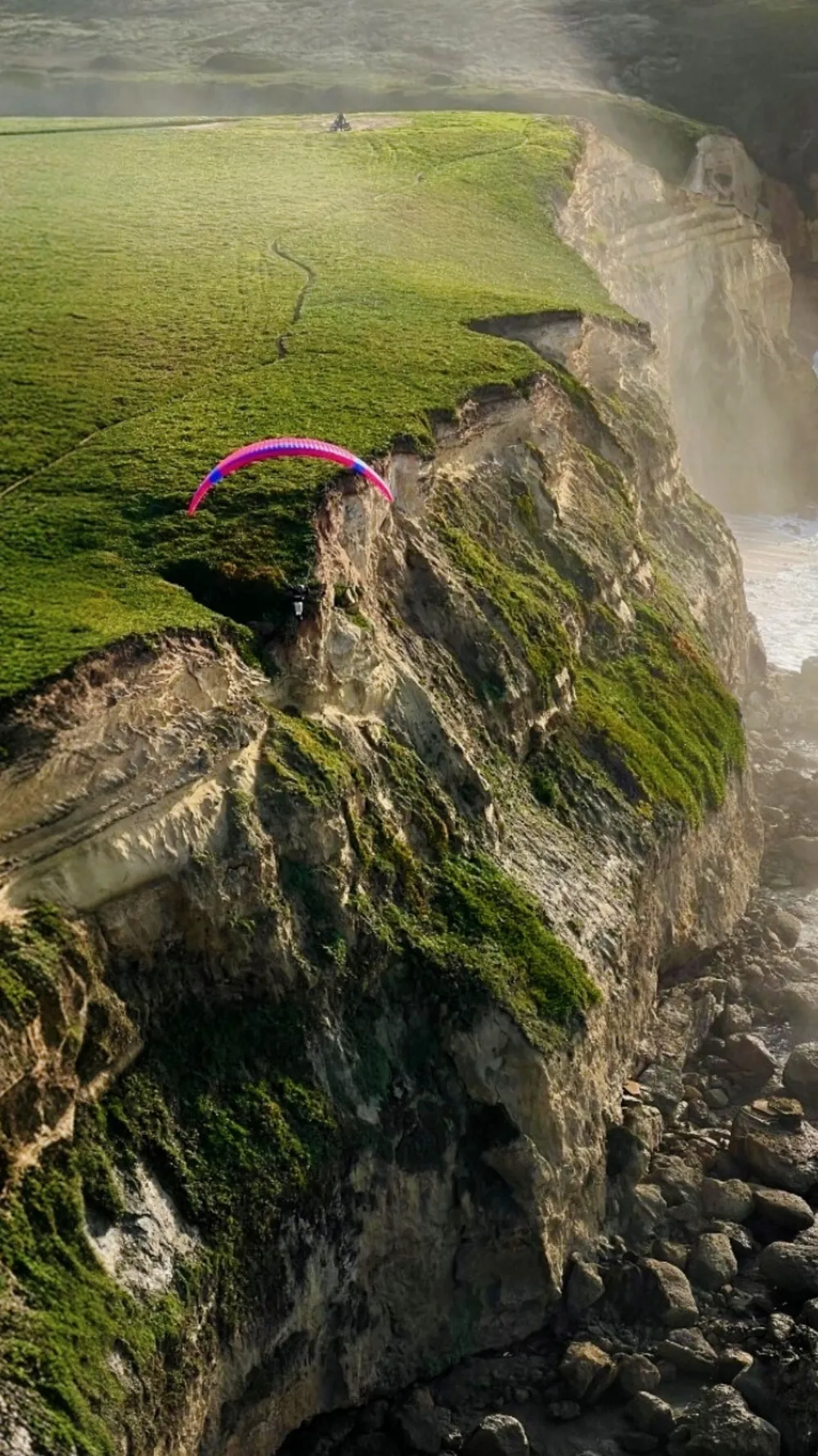 Parapente rosa a soarear rente à arriba verde da Praia das Bicas, com neblina do mar e rochas em baixo.