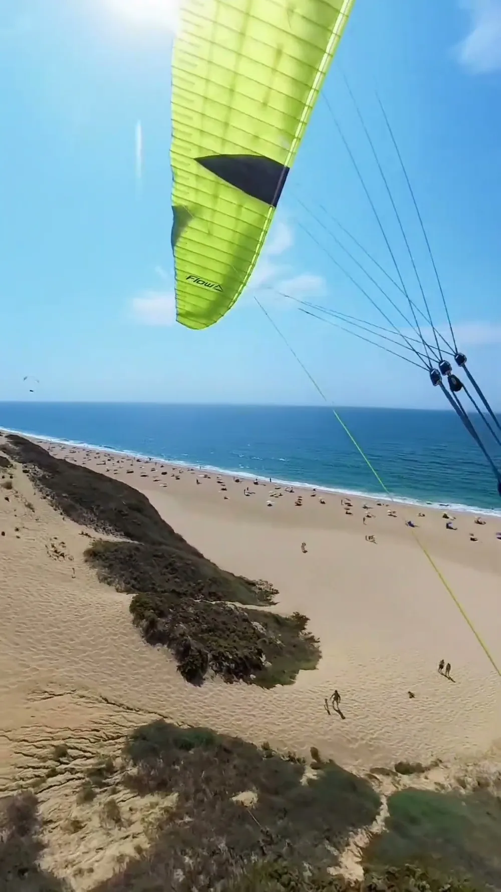 A vista do piloto sob uma asa Flow verde-fluorescente, a voar rente à arriba da Praia do Meco sobre um areal cheio de banhistas, com outra asa ao longe.